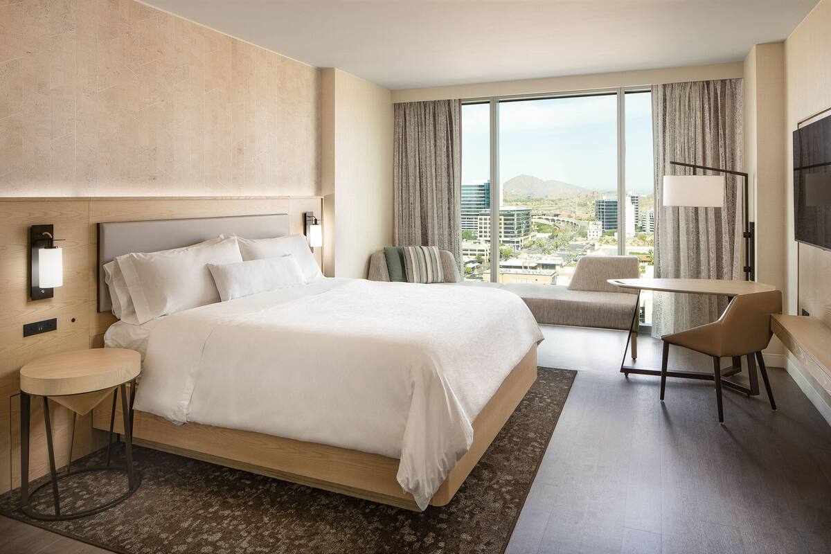 A modern hotel room with a large bed, a small table, a chair, a TV, and a view of the city.