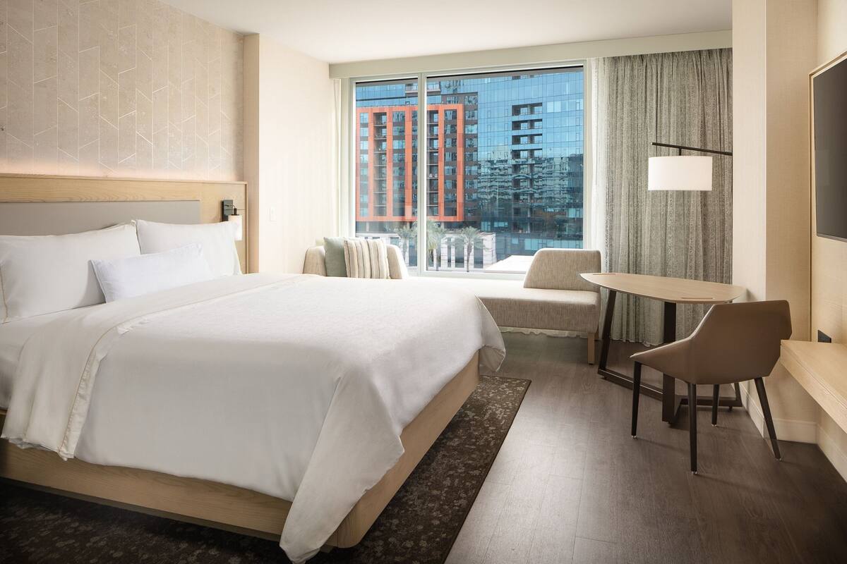 A hotel room with a large bed, a desk, a chair, and a view of a building through the window.