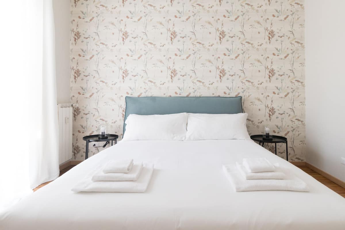 A neatly made bed with white linens and towels, a blue headboard, and floral wallpaper.