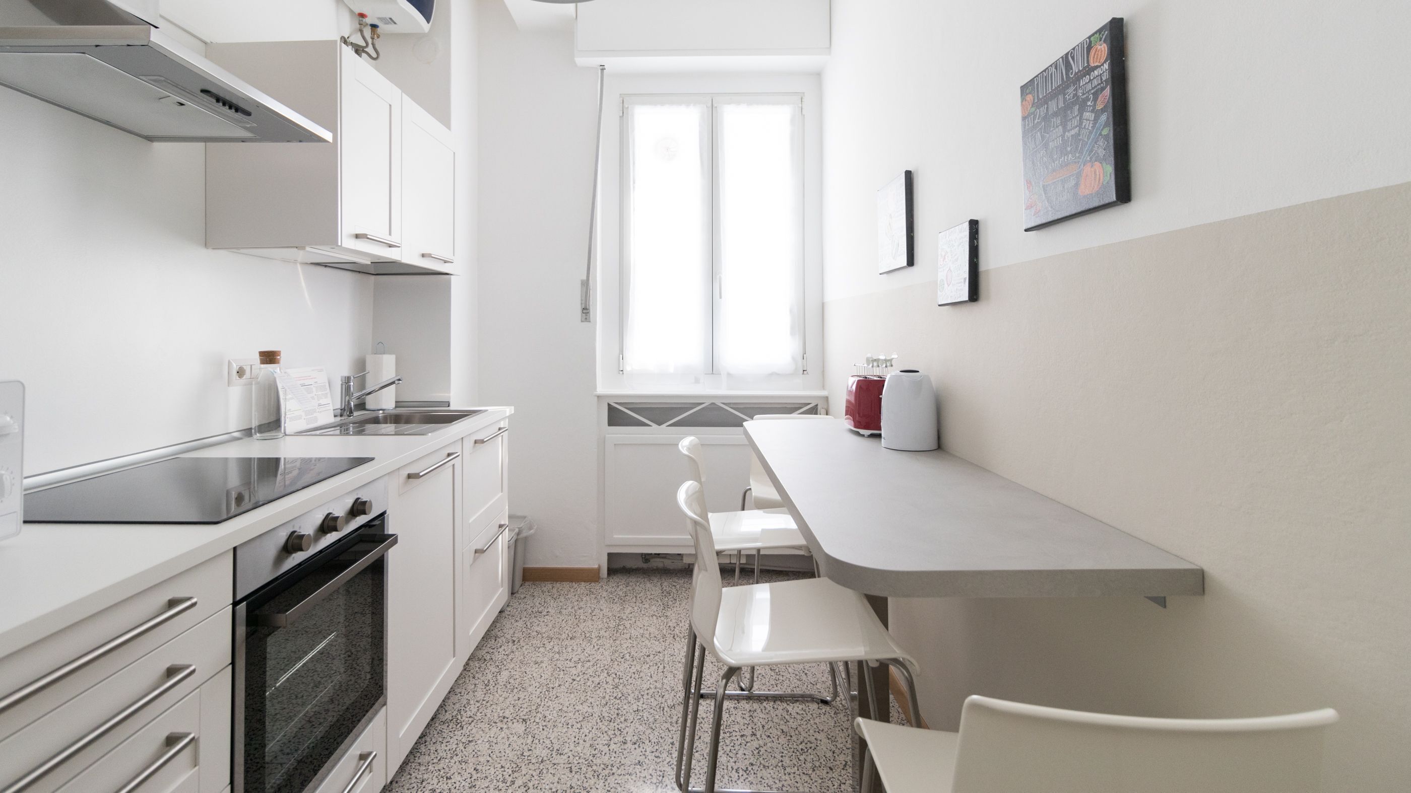 A compact kitchen with white cabinetry, a stainless steel sink, a built-in oven, and a window with sheer curtains.