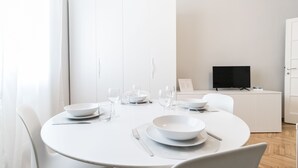 Een moderne eetruimte met een ronde tafel voor twee personen, witte stoelen en een flatscreen televisie op een standaard op de achtergrond.