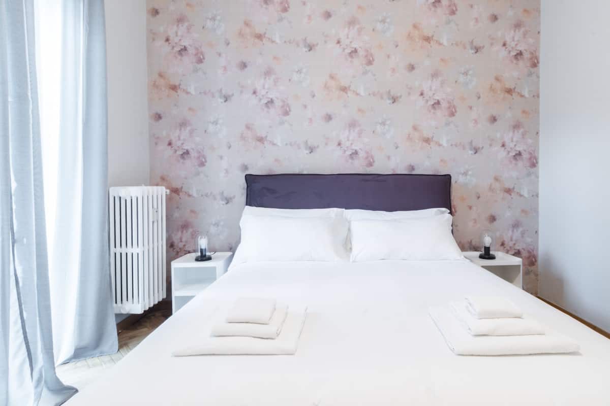 A neatly made bed with white linens and two pillows, a white bedside table with a lamp, and a radiator on the wall.
