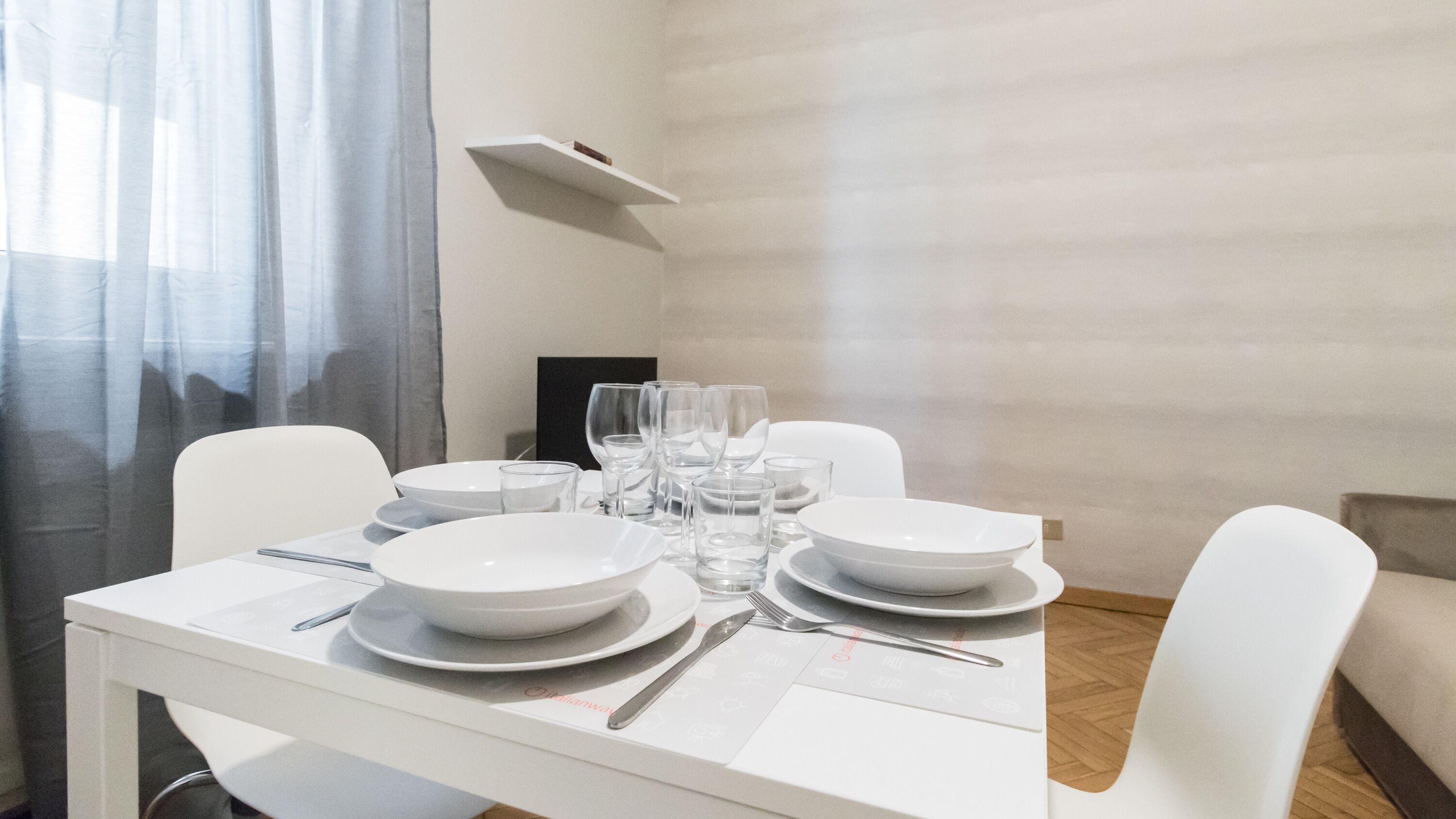 A dining table set with white plates, bowls, glasses, and cutlery in a modern room with a light-colored wall and a window with sheer curtains.