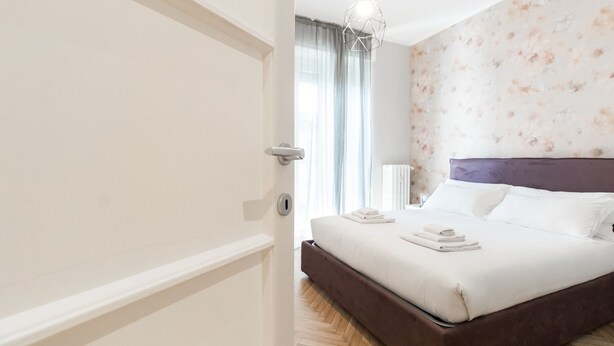 Een hotelkamer met een bed, nachtkastje, raam met gordijnen en een muur met een bloemenprint.