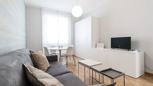 Een moderne, minimalistische woonkamer met een grijze bank, een kleine eettafel en een televisie op een wit kastje.