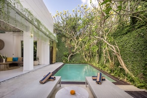 A modern outdoor pool area with lounge chairs, a small round table, and a lush green backdrop. - AV Villa Casabola (Seminyak)