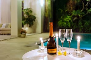 A table with a bottle of wine, two glasses, and candles in a well-lit indoor setting with a pool and greenery in the background. - AV Villa Casabola (Seminyak)