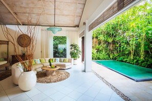A modern living room with a sofa, coffee table, and a large window overlooking a lush garden and pool. - AV Villa Casabola (Seminyak)