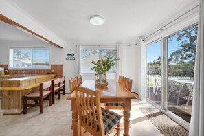A dining area with a wooden table and chairs, a kitchen area with a bar, and large windows with blinds.