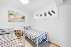 Une chambre avec deux lits, une fenêtre avec des stores, un tableau au mur et une petite table de chevet avec une lampe.