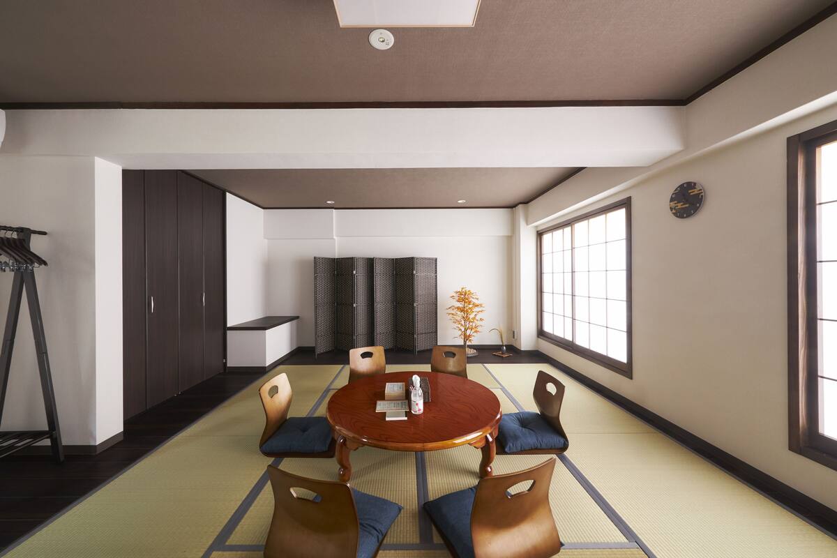A modern conference room with a round wooden table, six chairs, tatami mats, a bookshelf, and a large window.