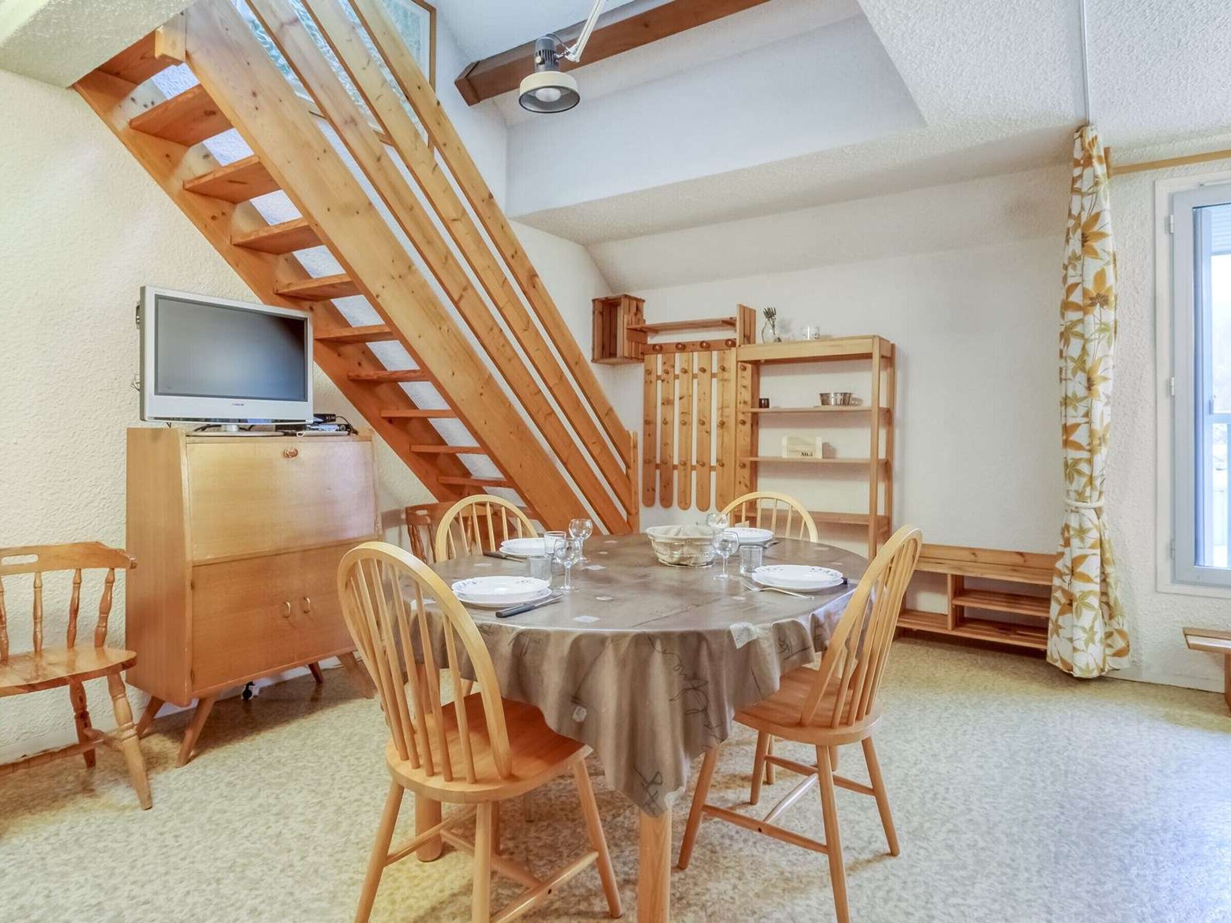 Un coin repas avec une table ronde, six chaises en bois, un buffet en bois, un téléviseur posé sur un meuble et un escalier en bois.
