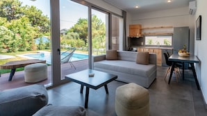 A modern living room with a sofa, ottomans, and a dining table. A kitchen with a sink and cabinets is visible through an open door. - SunBlue Villa Modern Private Pool (thasos)