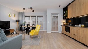 A modern living room with a sofa, a yellow chair, a dining area with a table and chairs, a kitchen with wooden cabinets, and a TV.
