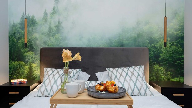 A neatly made bed with a tray of pastries and two cups, a vase with flowers, and a forest mural on the wall.