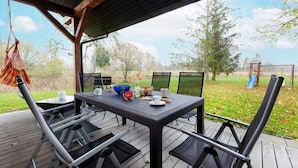 An outdoor patio with a table and chairs, a hanging chair, and a view of a grassy area with trees and a playground.
