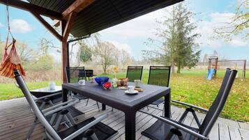 Une terrasse extérieure avec une table et des chaises, un fauteuil suspendu et une vue sur un espace verdoyant agrémenté d’arbres et d’une aire de jeux.