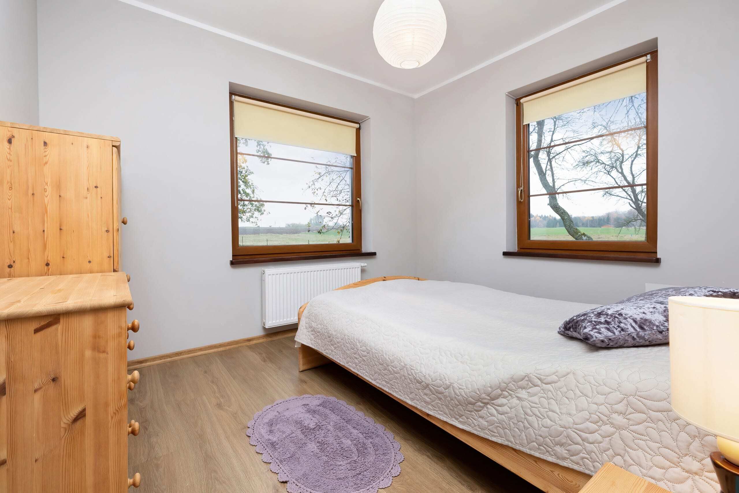 A bedroom with a bed, a wooden wardrobe, two windows with blinds, a radiator, and a purple rug.