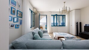 A modern bedroom with a large sofa, a bed with a patterned bedspread, a chandelier, and a view of the sea through a large window. - Villa Chryssi - Hilltop Escape w/ Endless Sea Views by etouri (Chania)