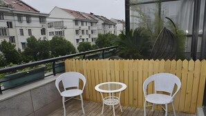 Een balkon met twee witte rieten stoelen en een klein rond tafeltje, met uitzicht op een woongebied met gebouwen en groen.
