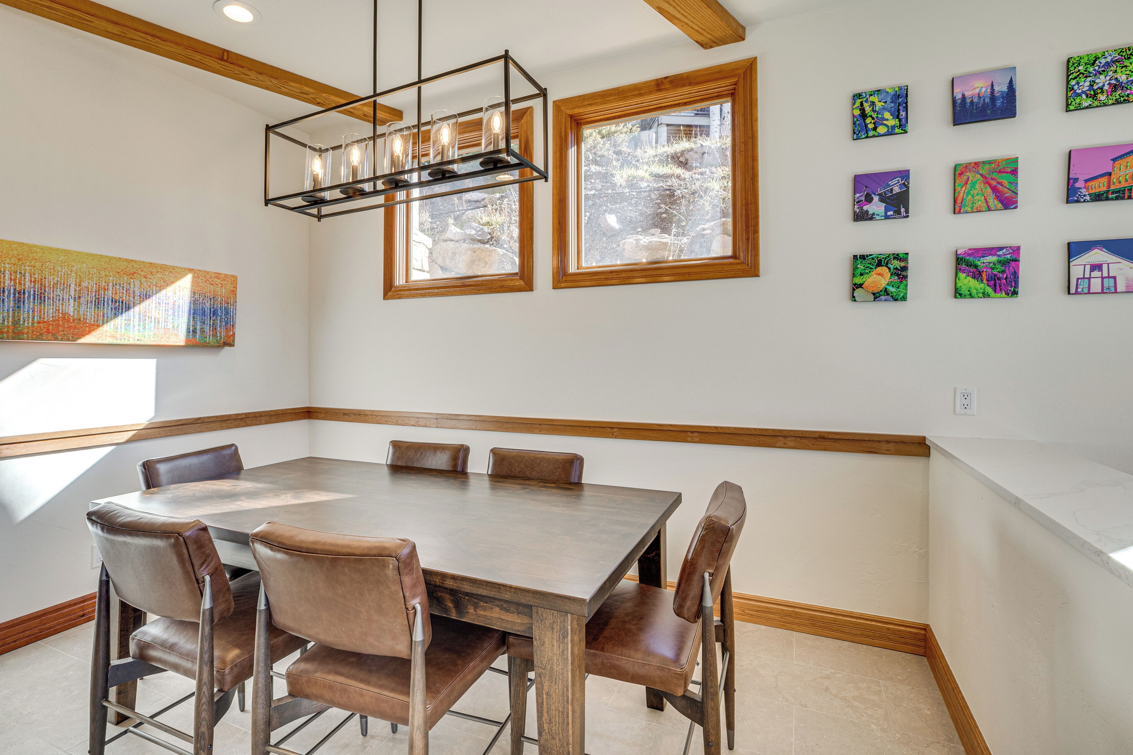A dining area with a large wooden table and leather chairs, surrounded by large windows and a modern chandelier.