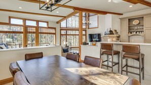 A spacious dining area with a large wooden table and leather chairs, a kitchen with bar stools, and large windows offering a view of the outdoors. - Luxurious Ski-In/Ski-Out Telluride Mountain Escape (Mountain Village)