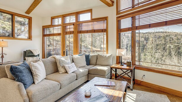 A cozy living room with a sectional sofa, wooden beams, large windows, and a view of a forest. - Luxurious Ski-In/Ski-Out Telluride Mountain Escape (Mountain Village)