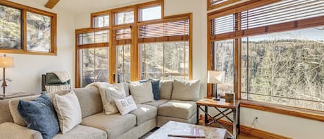 Una sala de estar acogedora con un sofá modular, vigas de madera, ventanas amplias y vistas a un bosque.
