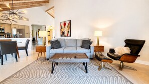 Una sala moderna con un sofá, una mesa de centro y sillas, una cocina al fondo y un cuadro en la pared.