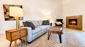 Uma sala de estar com sofá, mesa de centro, mesa lateral, luminária, poltrona e lareira.