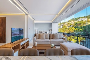 A modern living room with a sofa, coffee table, and TV, a balcony with a city view, and a dining area with chairs.