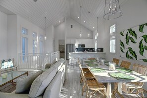 A spacious dining area with a long table and wicker chairs, a kitchen in the background, and a balcony with a railing.