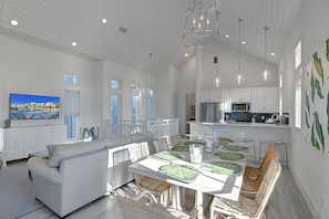 A spacious living area with a large TV, a dining table with chairs, and a kitchen in the background.