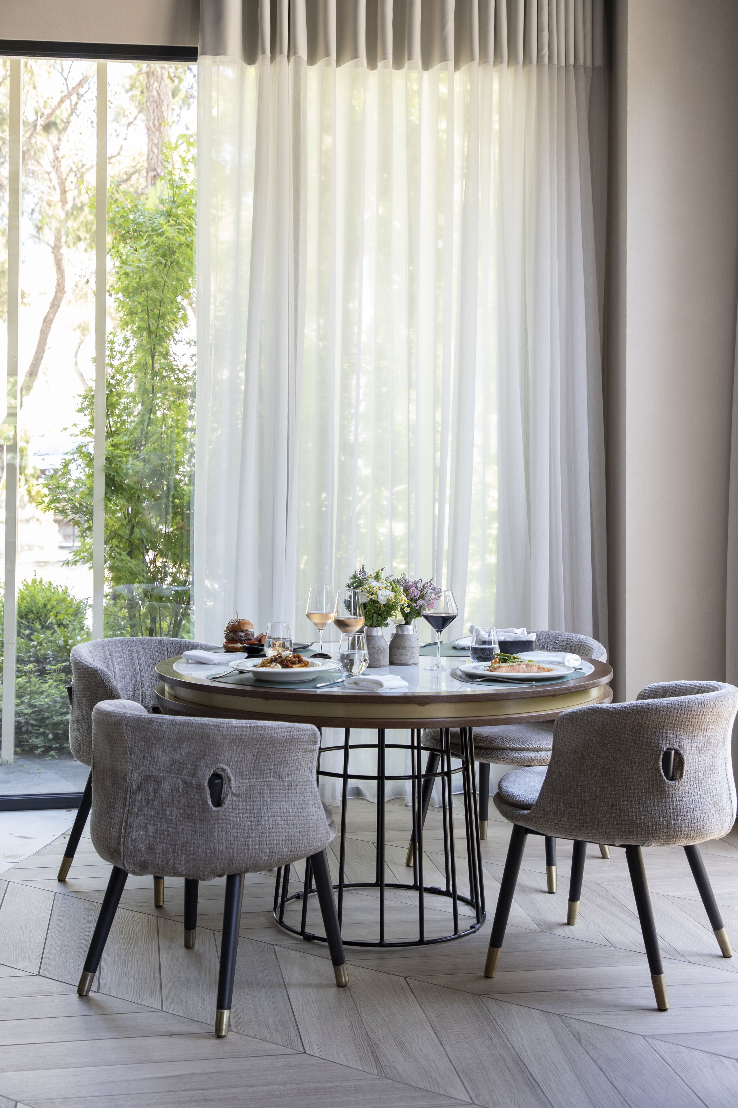 Un comedor con una mesa redonda, sillas tapizadas en gris y vistas a zonas verdes a través de cortinas traslúcidas.