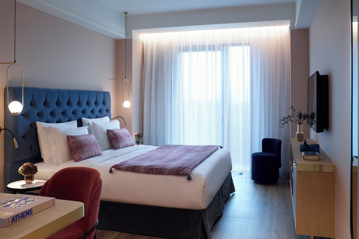 Una habitación de hotel moderna con una cama grande, un escritorio con un libro, una silla, un televisor y una ventana con cortinas ligeras.