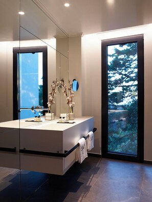 Une salle de bain moderne avec un grand miroir, un lavabo et une vue sur une forêt, visible à travers une porte en verre.