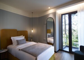 A modern hotel room with a large bed, a bedside table, a mirror, and a view of the outdoors through a large window.