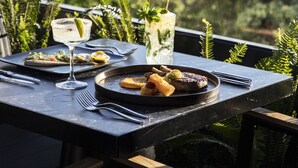 Una mesa de comedor con platos de comida, vasos y cubiertos, frente a un fondo de vegetación y un edificio lejano.