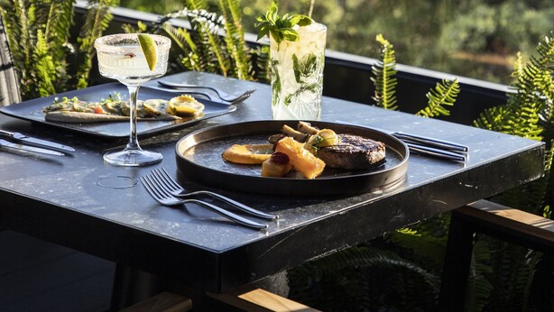 A dining table with plates of food, glasses, and cutlery, set against a backdrop of greenery and a distant building. - SAY Hotel (Kifisia)
