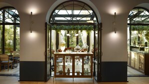 A room with a glass cabinet displaying various decorative items, a dining area with wicker chairs, and large windows with a view of greenery. - SAY Hotel (Kifisia)