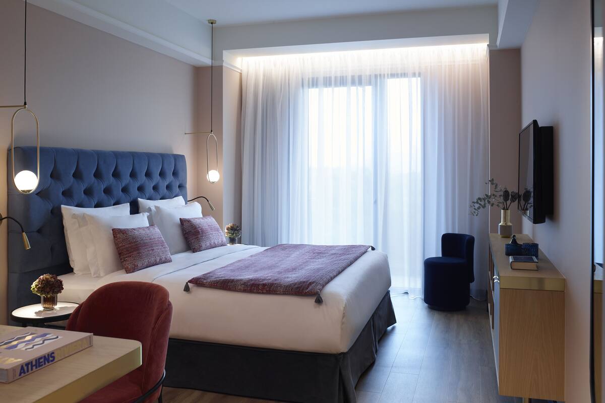 Moderni hotellihuone, jossa on suuri sänky, kirjoituspöytä, jossa on kirja, tuoli, televisio ja ikkuna, jossa on läpikuultavat verhot.