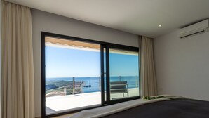 Une chambre dotée d’une grande porte-fenêtre coulissante donnant sur un balcon avec vue sur la mer et sur le paysage urbain au loin.