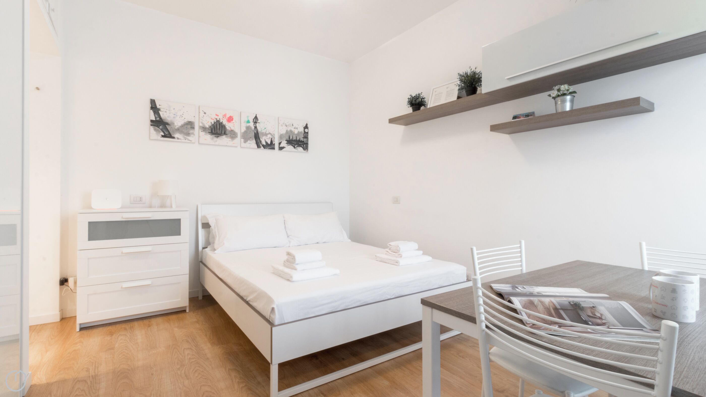 A modern bedroom with a bed, bedside table, and a desk with chairs. There are wall art pieces above the bed and shelves with plants on the wall.