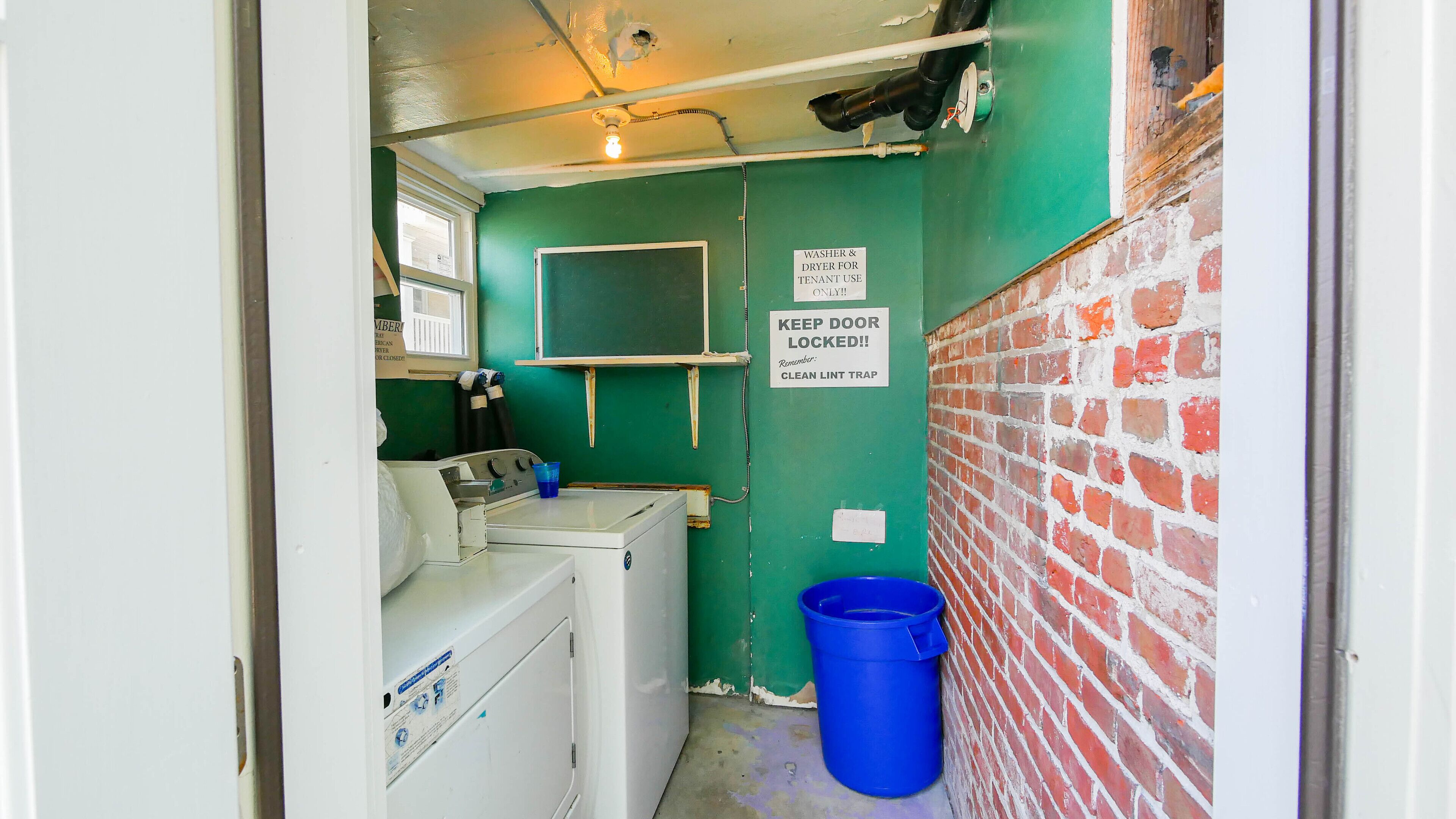A laundry room with a washing machine, a dryer, a blue bucket, and a sign instructing to keep the door locked.