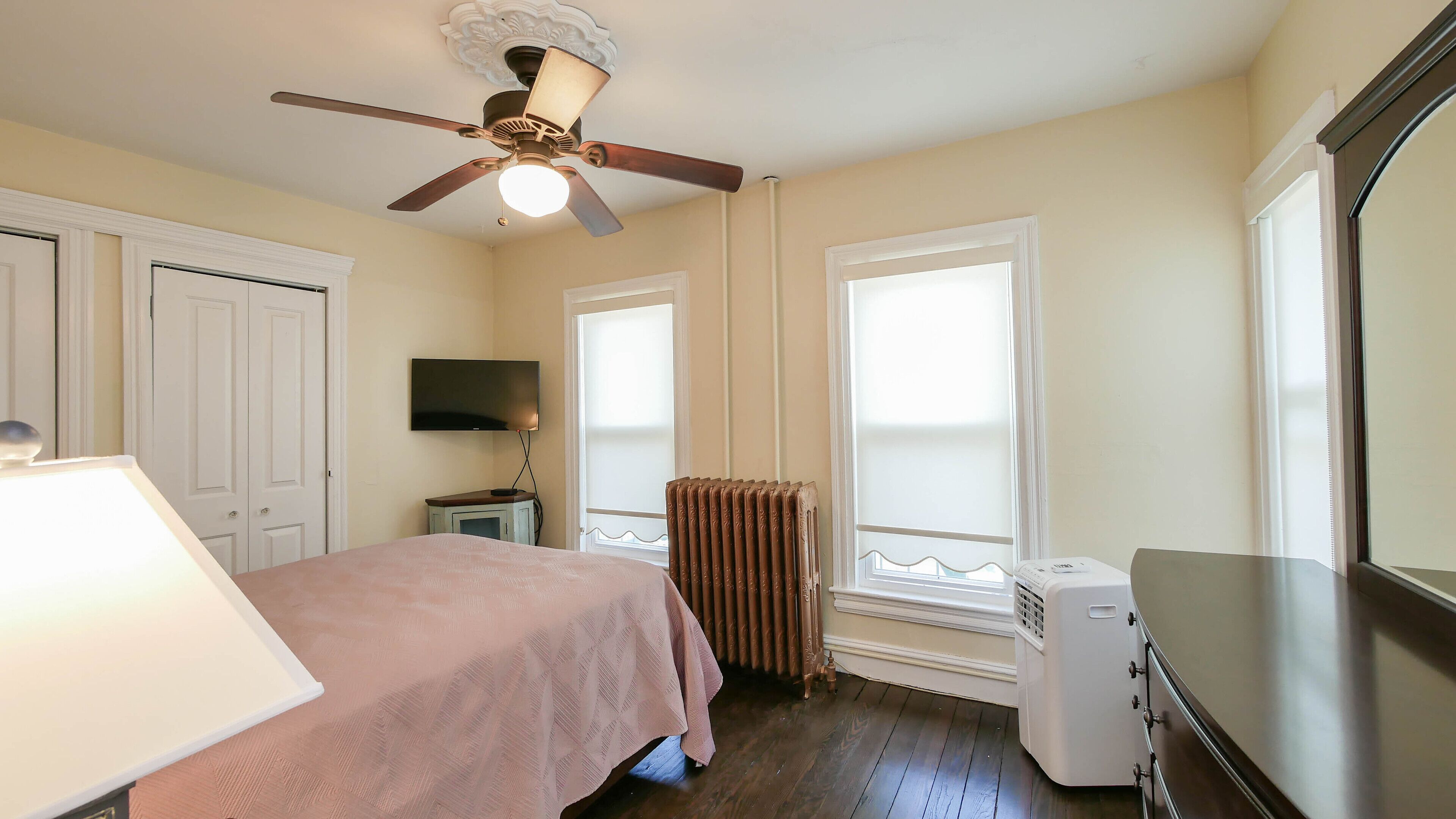 A bedroom with a bed, a ceiling fan, a radiator, a TV, and a dresser.