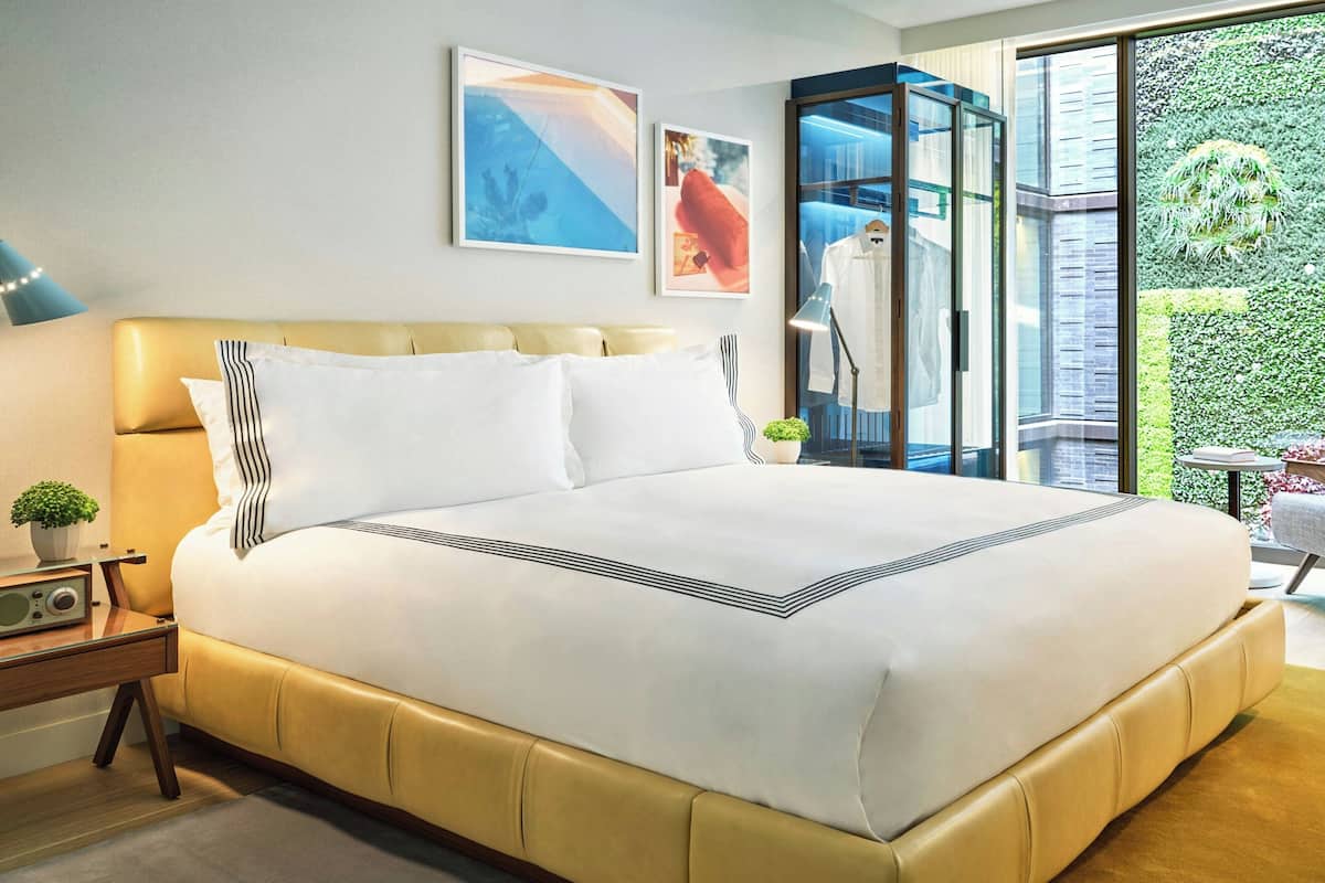 A modern hotel room with a large bed, a bedside table with a radio, and a view of a green garden through a glass door.