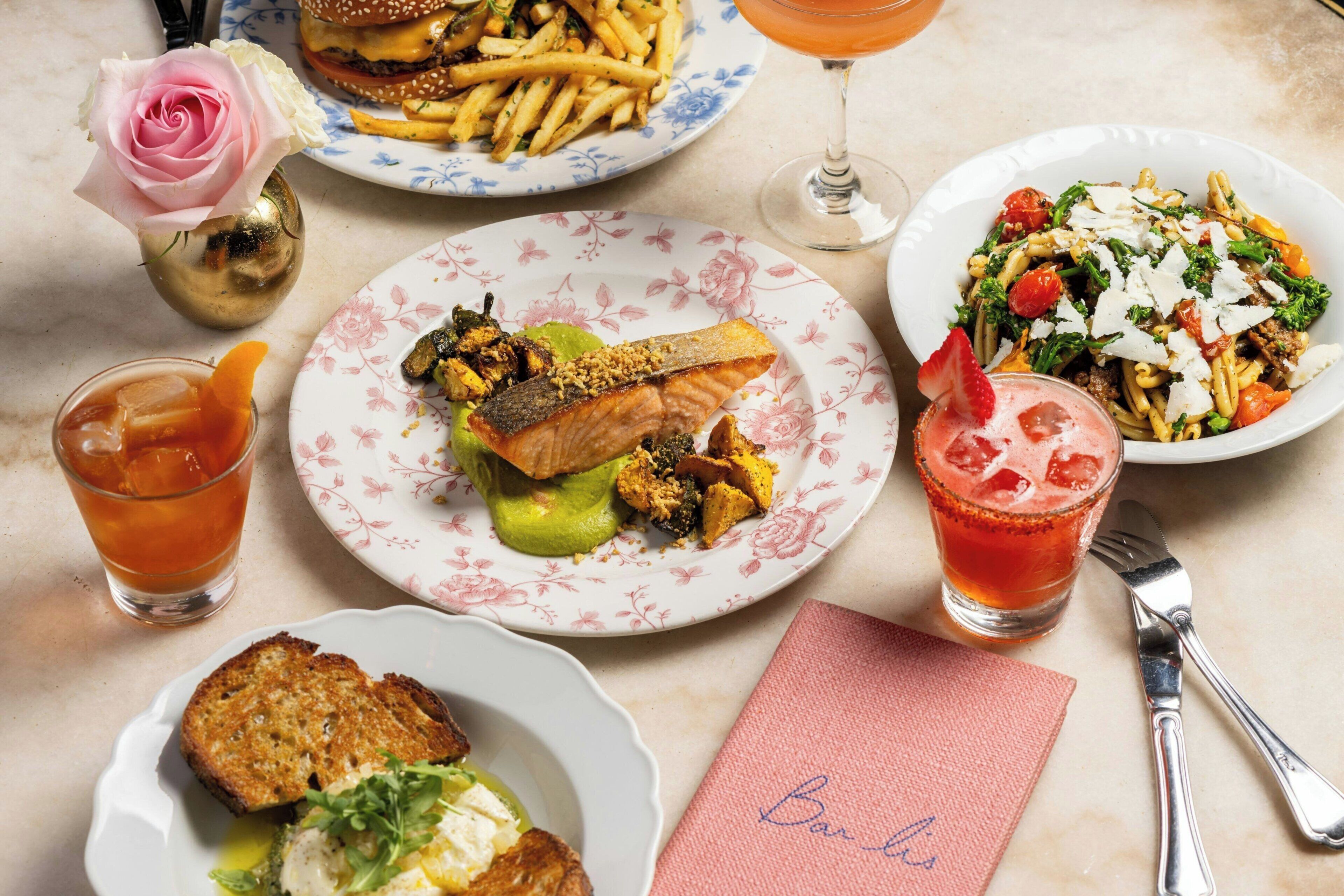 A table set with various dishes including a burger, fries, fish, pasta, and drinks, with a rose and a gold teapot visible.