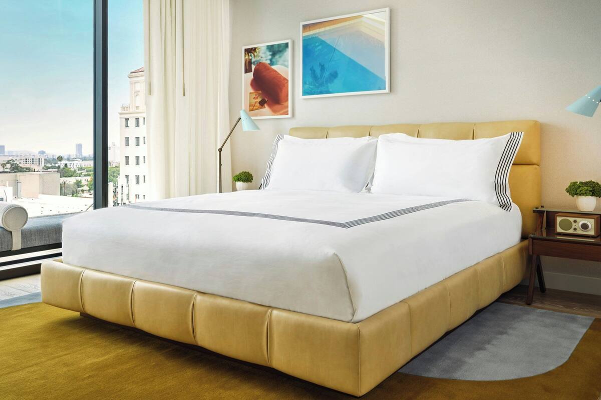A large bed with a beige upholstered headboard, white bedding, and a view of the city through a large window.