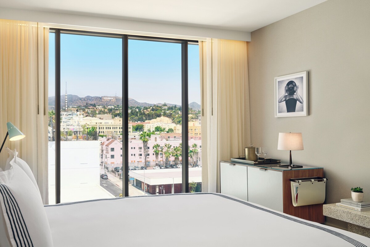 A hotel room with a large bed, a desk with a lamp, a view of a cityscape, and a framed picture on the wall.