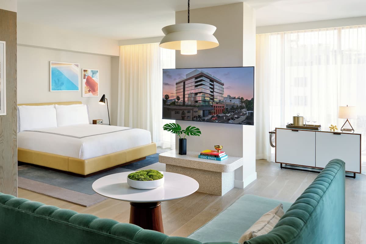 A modern hotel room with a bed, a round table with a plant, a green sofa, and a cityscape view.